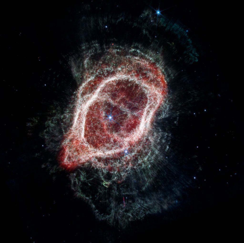 La vista de Webb de la Nebulosa del Anillo Sur aparece como un óvalo deforme que está ligeramente inclinado desde la parte superior izquierda hacia la parte inferior derecha. La imagen muestra una estrella en el centro. Un gran óvalo irregular rosa y rojo translúcido rodea las estrellas centrales.