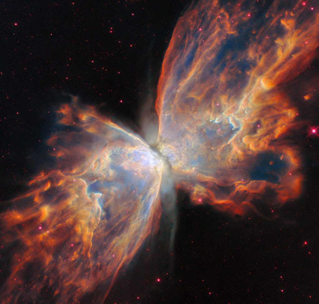 Una nebulosa planetaria llamada NGC 6302 o Nebulosa de la Mariposa. Una franja de polvo oscuro atraviesa el centro de la nebulosa y dos amplias nubes emergen a ambos lados de la franja de polvo, como las alas extendidas de una mariposa. La nebulosa tiene un aspecto grumoso y casi opaco, con algunas estrellas de fondo que se asoman a través del material nuboso. La nebulosa se presenta en diferentes tonos de crema, amarillo y naranja, siendo los colores más claros los que se encuentran más cerca del centro. El fondo del espacio es negro con unas pocas estrellas teñidas de rosa.